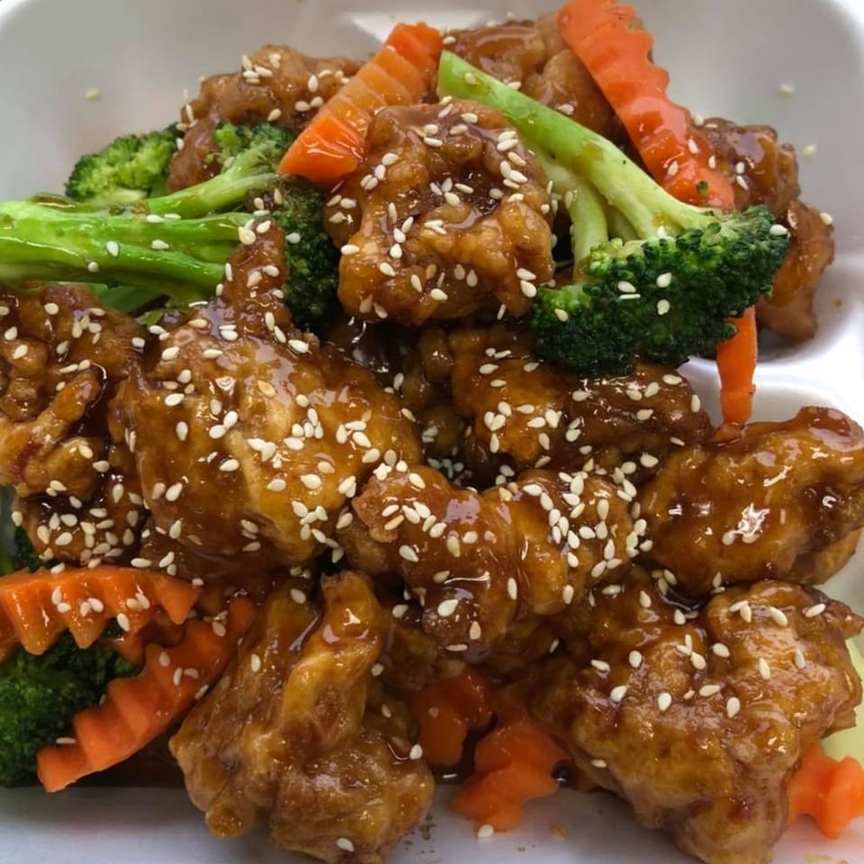 Sesame chicken with broccoli and carrots, glistening with sauce and sprinkled with sesame seeds.